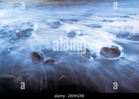 Lange Aufnahme von Surfwellen zwischen großen Kieselsteinen Stockfoto