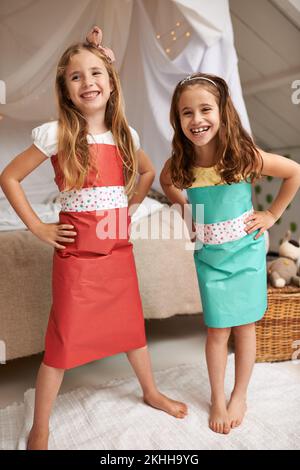 Schwestern machen die besten Freunde. Porträt von zwei kleinen Mädchen, die in den Kleidern posieren, die sie aus Papier gemacht haben. Stockfoto
