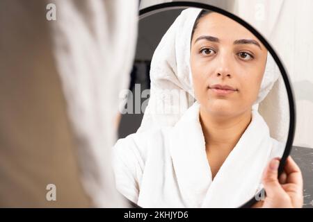 Eine schöne Frau, die nach dem Duschen im Spiegel steht, und ihr Haar in ein Handtuch gewickelt. Konzept der Morgenbetreuung. Körperhygiene, kosmetische Behandlungsverfahren. Stockfoto