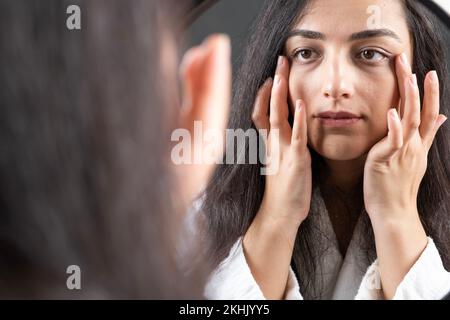 Berührendes Gesicht, hübsche brünette Frau, die spiegelverkehrt aussieht. Feuchtigkeitsspendende weiche Haut. Hautpflegekonzept. Fühlt sich zufrieden, Kosmetiker. Stockfoto