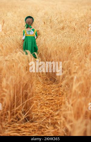 Kleines Mädchen im russischen nationalen Sarafan und ein Kokoschnik, der am Sommertag in einem goldenen Weizenfeld steht Stockfoto