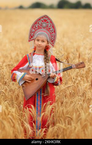 Kleines Mädchen im russischen nationalen Sarafan und ein Kokoschnik, der am Sommertag in einem goldenen Weizenfeld steht Stockfoto