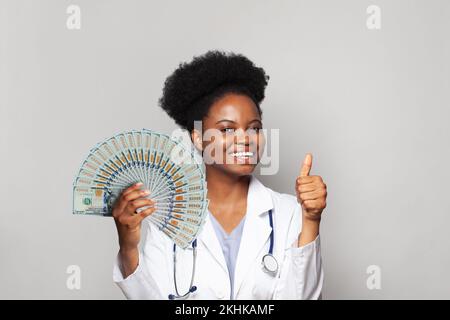 Erfolgreiche afroamerikanische Ärztin lächelt und hält Fans von Geld und Daumen nach oben auf weißem Hintergrund Stockfoto