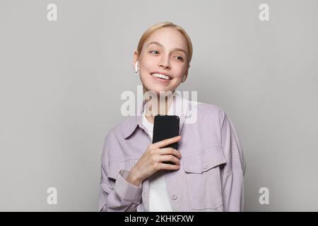 Glückliche junge Frau, die Musik oder Podcast mit Kopfhörern hört und das Smartphone auf grauem Hintergrund hält Stockfoto