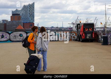 Zwei Touristen mit Koffer und Rucksack auf einer Aussichtsplattform im Hamburger Hafen am Landungsbrücken. Im Hintergrund die Elbphilharmonie. Stockfoto