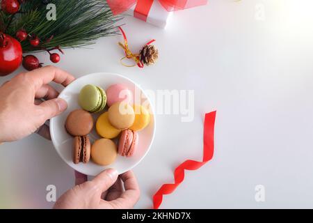 Handservierungs-Stapel französischer Makronen verschiedener Geschmacksrichtungen auf Keramikplatte auf weißem Tisch mit Weihnachtsdekoration. Draufsicht. Stockfoto