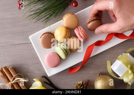 Mit der Hand holte man sich eine Schokoladenmakarone von einem Teller voller verschiedener Geschmacksrichtungen auf einem Holztisch mit Weihnachtsdekoration. Draufsicht. Stockfoto