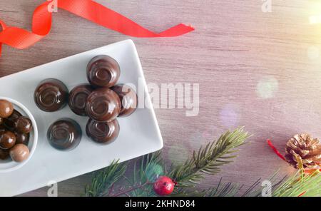 Details einer weißen Keramikplatte gefüllt mit dunklen Schokoladenbonbons auf einem Holztisch mit Weihnachtsdekorationen. Draufsicht. Stockfoto