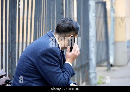Ein Mann in einem Business-Anzug, der mit dem Handy spricht und auf einer Bank sitzt. Das Konzept der zellulären Kommunikation auf der Straße der Stadt Stockfoto