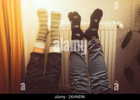 Ein Paar hält die Beine hoch und heizt die Füße in kalten Wohnungen drinnen auf der Heizung im Winter mit kuscheligen, stilvollen Wollsocken an. Valentinstag lustig zusammen warm f Stockfoto
