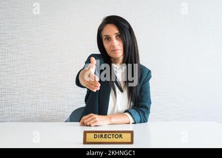 Die junge, brünette Geschäftsfrau im Büro hinter dem Schreibtisch gibt den Mitarbeitern aus der Sicht des Kunden die Hand für den Händedruck Stockfoto