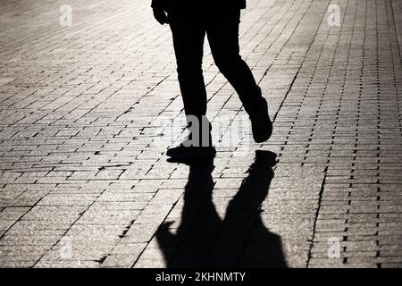 Schwarze Silhouette und Schatten eines einsamen Mannes, der auf einer Straße läuft. Männerbeine in Jeans und Turnschuhen, dramatisches Leben Stockfoto