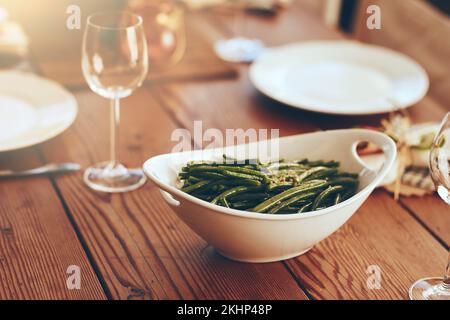 Thanksgiving, grüne Bohnen und Speisen mit Platz auf einem Esstisch für eine Feier. Party, Gericht und Abendessen mit einer Schüssel auf einem Holz Stockfoto