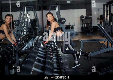 Schlankes Bodybuilder-Mädchen, hebt schwere Kurzhanteln, steht vor dem Spiegel, während es im Fitnessstudio trainiert. Sportkonzept, Fettverbrennung und ein gesundes Leben Stockfoto