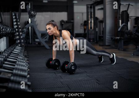 Schlankes Bodybuilder-Mädchen, hebt schwere Kurzhanteln, steht vor dem Spiegel, während es im Fitnessstudio trainiert. Sportkonzept, Fettverbrennung und ein gesundes Leben Stockfoto