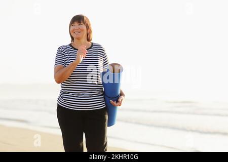 Eine Frau mittleren Alters hält eine Sportmatte und bereitet sich darauf vor, im Freien am Meeresstrand Yoga zu üben. Glückliche, reife, übergewichtige Frau, die an der Küste trainiert. Speicherplatz kopieren. Meditation, Yoga und Entspannungskonzept Stockfoto