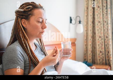 Frau, die im Bett sitzt und Glas Wasser und Pille in den Händen hält. Stockfoto
