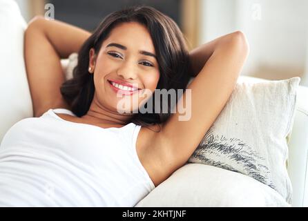 Porträt, entspannt und glücklich mit einer schwarzen Frau, die über ein Wochenende auf dem Sofa im Wohnzimmer ihres Hauses liegt. Gesicht, Lächeln und ruhen Sie sich mit einer jungen Frau aus Stockfoto