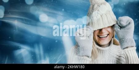 Lächelnde Frau in warmen Kleidern auf blauem Winterhintergrund. Versteck das Gesicht unter dem Strickmütze aus Wolle. Banner mit Kopierbereich Stockfoto