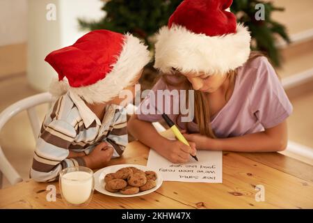 Kinder, Schreiben und Brief an den weihnachtsmann an einem Tisch mit Bruder und Schwester, die Wunschzettel für weihnachten mit Milch und Keksen planen. Kinder, Hoffnung und Aufzählung Stockfoto