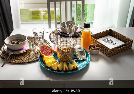 Gesundes Frühstück im Innenbereich mit köstlichem Haferbrei, Apfelscheiben, geschnittenen Grapefruits, Haselnüssen und Orangensaft auf einem weißen Holztisch an einem sonnigen Fenster Stockfoto