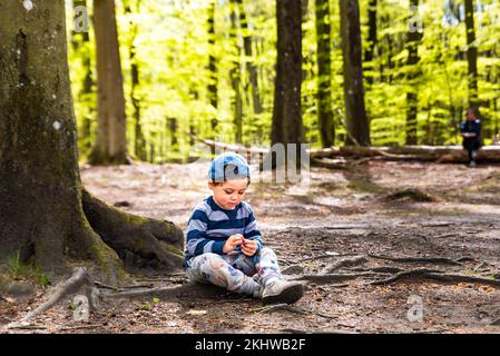 Ein Junge, der auf Baumwurzeln sitzt, ruht im Schatten eines Waldes. 5 Jahre altes Kind, das sich im Wald entspannt und dabei die Umgebung beobachtet Stockfoto