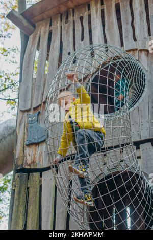 Ein Kind klettert an einem heißen Sommertag in einem Park auf einem Spielplatz auf ein Berggitter. Kinderspielplatz in einem öffentlichen Park, Unterhaltung und Erholung Stockfoto