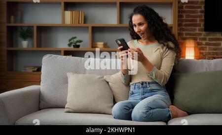 30s Hispanische Latina Weiße Frau Frau, die zu Hause auf der Couch sitzt und mit dem Handy spielt Online-Wetten Sieg hat Rabatt Stockfoto