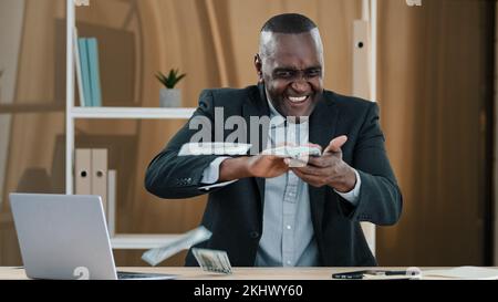 Glücklicher erfolgreicher alter afroamerikanischer Geschäftsmann älterer reifer Mann Arbeiter Investor im Büro, der am Tisch sitzt und Fan des Geldes hält, das Dollar wirft Stockfoto