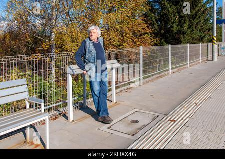 Senior lehnt sich auf eine schlanke Bank und wartet auf einen Zug auf einem Bahnsteig Stockfoto