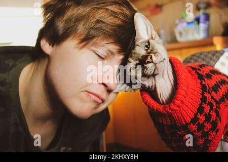 Eine Frau, die Katze liebt. Sie reibt ihren Kopf an den Kopf der Katze. Die beliebte kanadische Sphynx-Rasse. Geliebte und kahlköpfige Katze in rotem Pulli. Menschliches Haustier - Loyalität, Affe Stockfoto