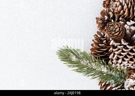 Weihnachtlicher Hintergrund mit Kiefernzapfen und einem Zweig mit grünen Nadeln, die mit Schnee bedeckt sind, isoliert auf silberweißem Hintergrund mit Kopierbereich. Stockfoto