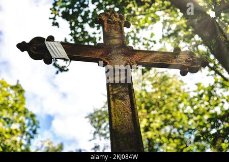 Ein rostiges Metallkreuz mit einer Gesichtsmaske, die auf einem alten Friedhof hängt. Schöne Sonnenstrahlen erhellen es. Stockfoto