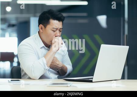 Ein junger asiatischer Freiberufler, ein Büroangestellter, wurde bei der Arbeit krank. Er sitzt am Tisch und hustet, bedeckt seinen Mund mit der Hand, hält seine Brust, fühlt sich schlecht. Stockfoto