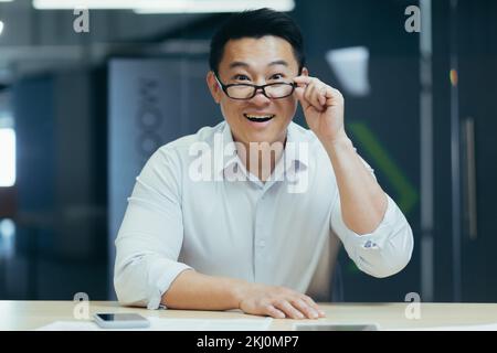 Porträt eines glücklichen jungen Asiaten, der im Büro am Tisch sitzt, mit Telefon und Dokumenten, mit Brille in der Hand, hinunter, lächelnd, in die Kamera schaut. Stockfoto