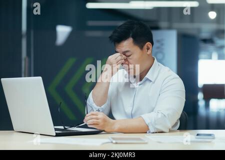 Ein müder, junger asiatischer Geschäftsmann. Hält seinen Kopf, fühlt Müdigkeit und Spannung in seinem Kopf und Körper, ist abgehauen und hält seine Brille. Er sitzt mit einem Laptop im Büro und braucht Ruhe. Stockfoto