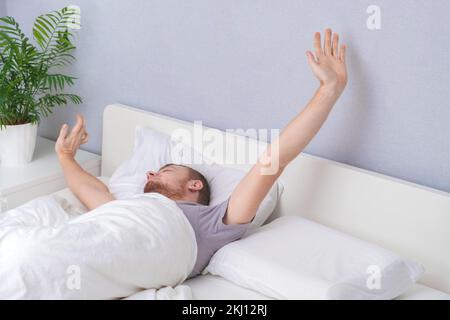 Ein glücklicher Mann wacht morgens nach dem Schlafen auf. Ein Mann ist nach dem Schlafen wach. Das faule Erwachen eines Mannes am Wochenende. Den Körper nach einem gesunden Schlaf aufwärmen. Stockfoto