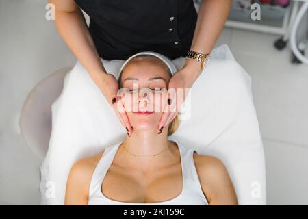 Blonde, hübsche und junge weiße Frau, die eine Massage im Gesicht bekommt, entspannend und Hautpflege in der Kosmetikklinik. Hochwertiges Foto Stockfoto