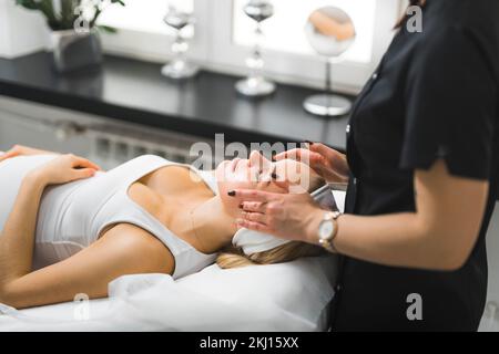 Eine Kosmetikerin in schwarzer Uniform, die eine Gesichtsmassage für ihre schwangere Klientin macht, ein mittelgroßes Schönheitszentrum mit Seitenblick. Hochwertiges Foto Stockfoto