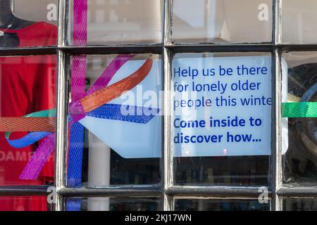 Melden Sie sich im Age UK Charity Shop „Helfen Sie uns, diesen Winter für einsame ältere Menschen da zu sein. Komm rein und entdecke, wie...“, England, Großbritannien, November 2022 Stockfoto