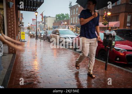 Ein Mann mit Schirm geht durch eine Regendusche in der Einkaufsstraße und geht an einem Mädchen vorbei, das mit seinen Armen mit den Regentropfen spielt Stockfoto