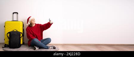 Eine junge Frau mit Koffer und Rucksack, die einen weihnachtsmannmütze trägt, benutzt ihr Smartphone, um sich auf einen Flug ins Ausland für das Wochenende vorzubereiten. Weihnachtstour Stockfoto