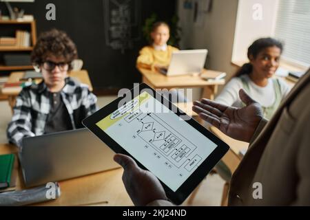 Nahaufnahme eines afroamerikanischen Lehrers, der ein digitales Tablet verwendet, mit Diagrammen auf dem Bildschirm, während er im Unterricht mit Kindern spricht Stockfoto