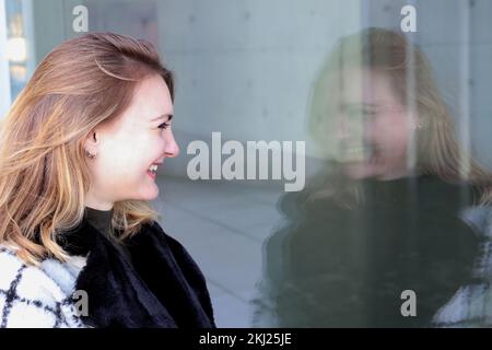 Eine junge Frau steht draußen und sieht ihr lachendes Spiegelbild am Fenster Stockfoto