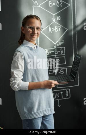 Porträt eines Schulmädchens mit Brille, das vor der Kamera lächelt, mit einem Laptop an der Tafel steht und IT-Technologie studiert Stockfoto