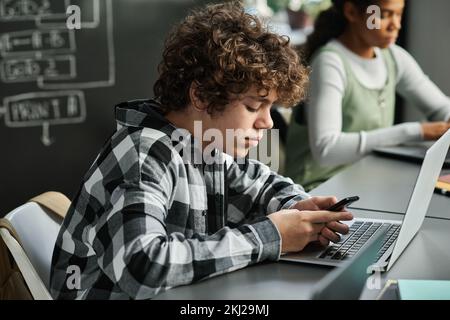 Der Schuljunge sitzt am Schreibtisch mit einem Laptop und benutzt sein Smartphone während des Unterrichts Stockfoto