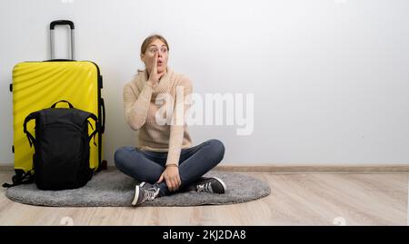 Eine Reisende mit Koffer im Pullover, die reisebereit ist, schreiend und mit Handfläche nahe dem geöffneten Mund. Passagier reist am Wochenende ins Ausland Stockfoto
