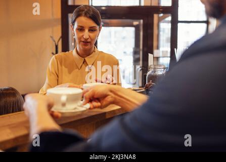 Männlicher Kellner, der einer Frau in der Cafeteria heißen Kaffee gibt. Ein Mann, der hinter der Theke steht und einer Frau in einem Café einen Cappuccino gibt. Porträt eines fröhlichen Stockfoto