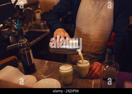 Der Barista gießt Milch in die Gläser auf dem Holztisch im Café. Zubereitung eines leckeren Kaffeegetränks mit Milch. Ein Mann hält einen Milchkrug aus Metall in der Hand Stockfoto