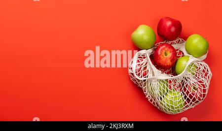Rote und grüne Äpfel im Netzbeutel auf rotem Hintergrund. Flach liegend mit Kopierbereich Stockfoto